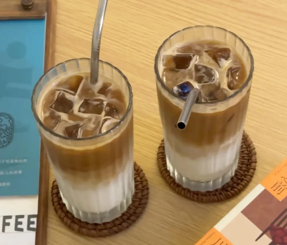 Q咖啡的兩杯冰拿鐵，牛奶與咖啡分層分明，裝在直紋玻璃杯裡放在藤編杯墊上